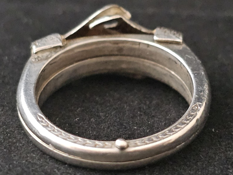 May include: Close-up of a silver ring with a unique design. The ring features a raised, architectural element and intricate detailing around the band. The ring is made of a shiny, metallic material, possibly sterling silver.