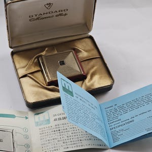 以下が含まれることがあります： 黒色のケースと青い説明書が付いた、ゴールドとレッドのヴィンテージなポケットラジオ。ラジオには「STANDARD」と「Miniature」の文字がケースに印刷されています。説明書には「TUNING IN」と「EARPHONE」の文字が印刷されています。