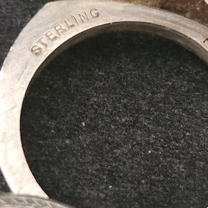 May include: Close-up of a silver ring with the word "STERLING" engraved on the band. The ring has a circular design with a dark background.