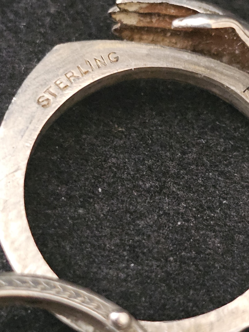 May include: Close-up of a silver ring with the word "STERLING" engraved on the band. The ring has a circular design with a dark background.
