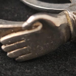 May include: A silver-toned bracelet with a hand-shaped design. The hand is detailed with visible fingers and a textured surface. The bracelet's design includes floral or leaf-like patterns near the wrist area. The background is a dark, possibly black, surface.