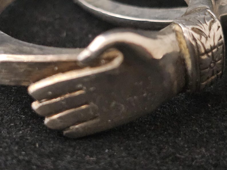 May include: A silver-toned bracelet with a hand-shaped design. The hand is detailed with visible fingers and a textured surface. The bracelet's design includes floral or leaf-like patterns near the wrist area. The background is a dark, possibly black, surface.
