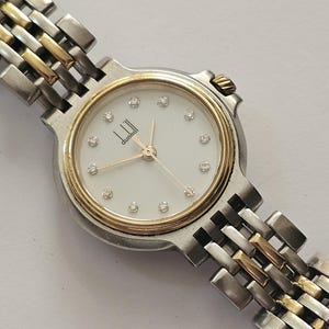 May include: A two-tone wristwatch with a silver and gold-coloured metal link bracelet. The round watch face features a white dial with gold-coloured hands, small sparkling stone hour markers, and the brand name dunhill printed in black text.