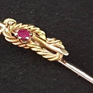 May include: A gold-toned tie tack featuring a red gemstone set in the centre. The tack is shaped like a knot with an intricate design.