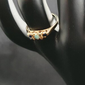 K&ouml;nnte beinhalten: Ein goldener Ring mit einem hellblauen Opal und zwei dunkelblauen Edelsteinen, die in einem floralen Design eingefasst sind.