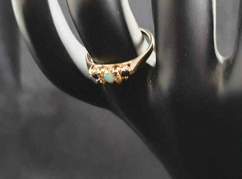 K&ouml;nnte beinhalten: Ein goldener Ring mit einem hellblauen Opal und zwei dunkelblauen Edelsteinen, die in einem floralen Design eingefasst sind.