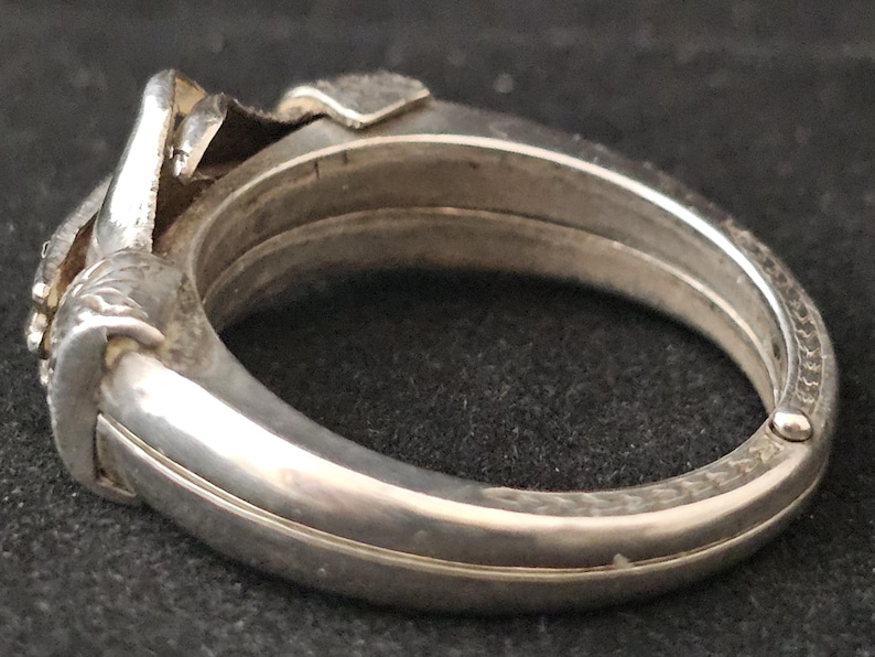 May include: A silver-toned hinged bangle bracelet. The bracelet has a detailed design on the clasp and decorative elements. The bracelet is made of metal and has a polished finish. The bracelet is a jewelry item.