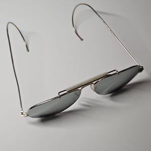 Pu&ograve; includere: Occhiali da sole con montatura argentata e lenti a specchio. Gli occhiali presentano un design sottile e curvo in filo di ferro per le aste e un ponte bianco. Le lenti sono di forma ovale e riflettono l'ambiente.