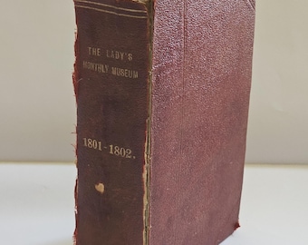 Puede incluir: Un libro antiguo de color granate, titulado "THE LADY'S MONTHLY MUSEUM", con los años 1801-1802 impresos en el lomo. El libro tiene una cubierta texturizada y bordes envejecidos, lo que indica su antigüedad.