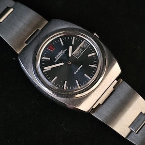 May include: A silver Omega wristwatch with a black face and a silver metal strap. The watch face has white markings and a date window that reads "TUE 27". The watch face also has the text "OMEGA" and "MEGAQUARTZ 38 KHZ" printed on it. The watch face also has the text "Geneve" printed on it.
