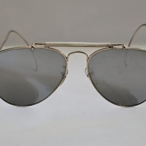 Pu&ograve; includere: Occhiali da sole in stile aviatore con lenti a specchio argentate e montatura in metallo argentato. Gli occhiali hanno un ponte bianco e aste sottili e curve. Le lenti sono a goccia.