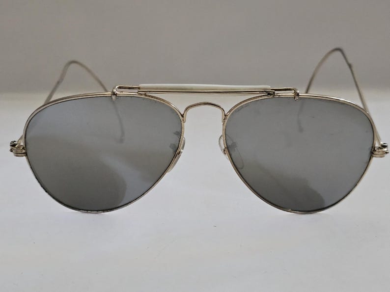 Pu&ograve; includere: Occhiali da sole in stile aviatore con lenti a specchio argentate e montatura in metallo argentato. Gli occhiali hanno un ponte bianco e aste sottili e curve. Le lenti sono a goccia.