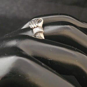 May include: A silver ring featuring a detailed fist design. The ring's fist is raised, with intricate detailing on the knuckles and a band adorned with small, sparkling accents. The ring is set against a dark background.