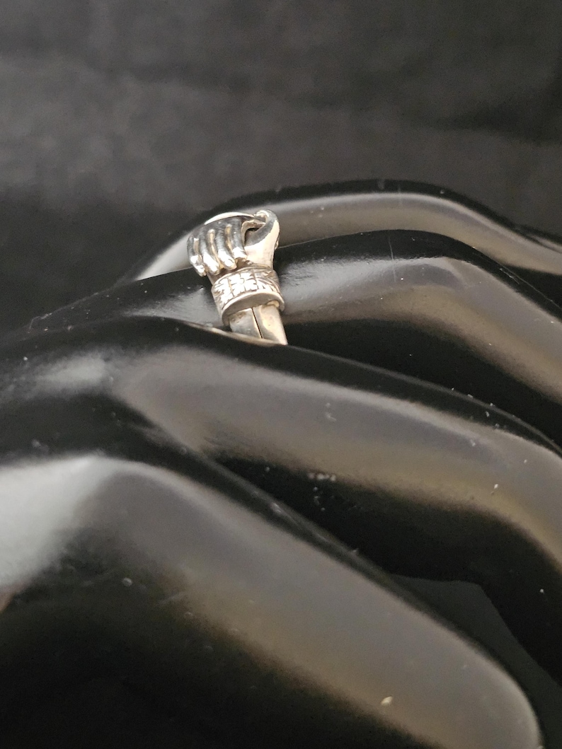 May include: A silver ring featuring a detailed fist design. The ring's fist is raised, with intricate detailing on the knuckles and a band adorned with small, sparkling accents. The ring is set against a dark background.