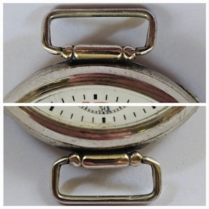 Op de afbeelding: Een zilveren zakhorloge met een witte wijzerplaat en zwarte markeringen. Het horloge heeft een unieke ovale vorm en een scharniergesp.