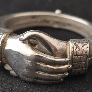 May include: A silver ring featuring two clasped hands, symbolizing unity. The ring has detailed carvings on the band and around the hands. The metal has a polished finish, reflecting light. The ring's design is intricate and symbolic.