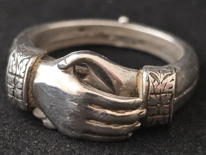 May include: A silver ring featuring two clasped hands, symbolizing unity. The ring has detailed carvings on the band and around the hands. The metal has a polished finish, reflecting light. The ring's design is intricate and symbolic.