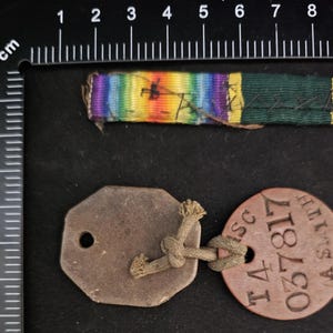 May include: A brown leather dog tag with the text "T4 037817 A SMITH" and a smaller, octagonal, brown leather tag with a hole in the centre. A multi-coloured ribbon with a green stripe is also visible.