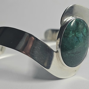 Puede incluir: Un brazalete de plata pulida con una banda ancha y curva que se estrecha hacia los extremos. Una gran piedra ovalada de color verde oscuro con sutiles vetas negras está engastada de forma prominente en un lado del marco de metal.