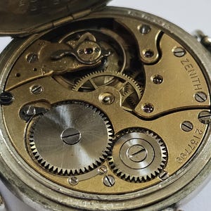 Op de afbeelding: Close-up van de binnenkant van een zilveren zakhorloge met een gouden wijzerplaat. Het horloge is open en de ingewikkelde tandwielen zijn zichtbaar. De wijzerplaat van het horloge heeft het woord "ZENITH" erop gedrukt en het nummer "667212".