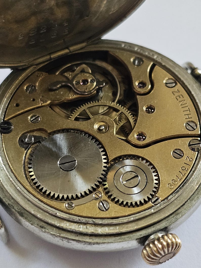Op de afbeelding: Close-up van de binnenkant van een zilveren zakhorloge met een gouden wijzerplaat. Het horloge is open en de ingewikkelde tandwielen zijn zichtbaar. De wijzerplaat van het horloge heeft het woord "ZENITH" erop gedrukt en het nummer "667212".