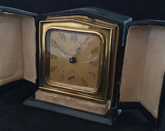 Könnte beinhalten: Ein goldfarbener Wecker mit schwarzen Zeigern und römischen Ziffern auf dem Zifferblatt, in einem schwarzen Lederetui mit Samtfütterung.