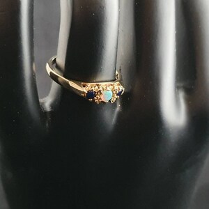 K&ouml;nnte beinhalten: Ein goldener Ring mit einem hellblauen Opal und zwei dunkelblauen Edelsteinen, die in einem filigranen, verzierten Design gefasst sind.