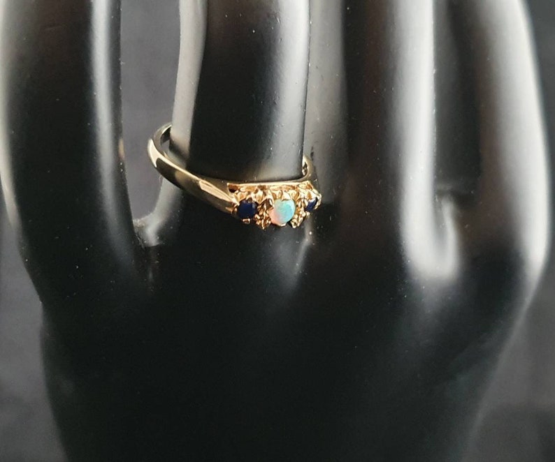 K&ouml;nnte beinhalten: Ein goldener Ring mit einem hellblauen Opal und zwei dunkelblauen Edelsteinen, die in einem filigranen, verzierten Design gefasst sind.