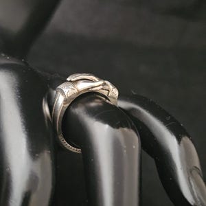May include: A silver ring with a detailed design. The ring features a sculpted hand motif, with intricate detailing on the fingers and palm. The band has a textured pattern. The ring is displayed on a black surface.