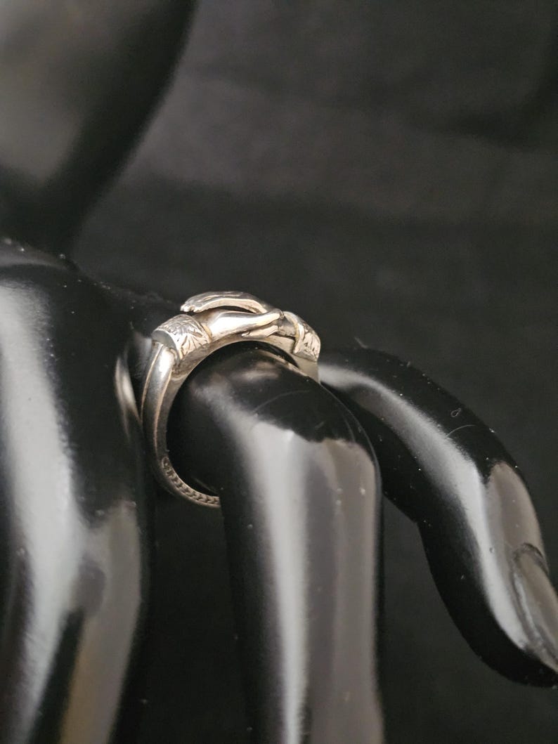 May include: A silver ring with a detailed design. The ring features a sculpted hand motif, with intricate detailing on the fingers and palm. The band has a textured pattern. The ring is displayed on a black surface.