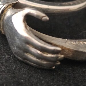 May include: Close-up of a silver ring with a hand design. The ring is made of sterling silver and features a detailed depiction of a hand holding a ring. The word "STERLING" is visible on the ring's band. The ring is on a dark surface.