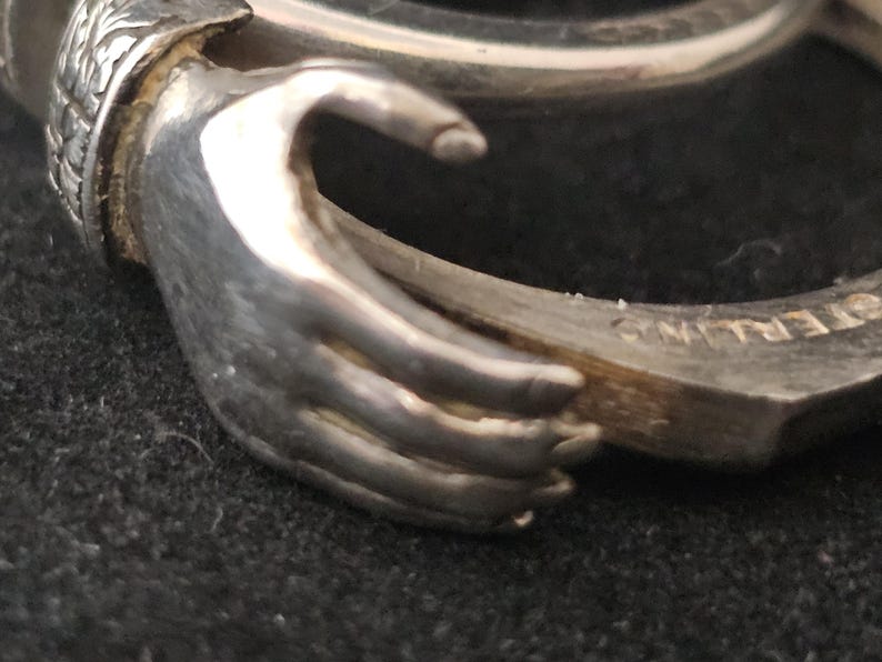 May include: Close-up of a silver ring with a hand design. The ring is made of sterling silver and features a detailed depiction of a hand holding a ring. The word "STERLING" is visible on the ring's band. The ring is on a dark surface.