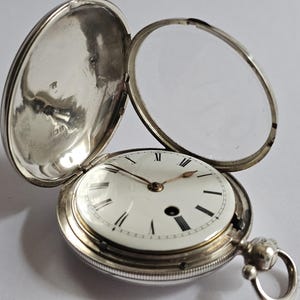 Könnte beinhalten: Eine offene antike Taschenuhr mit silbernem Gehäuse und weißem Zifferblatt. Das Zifferblatt der Uhr hat schwarze römische Ziffern und Zeiger. Der Deckel der Uhr ist geöffnet und gibt das Innenleben frei. Die Uhr hat eine Schlaufe für eine Kette.