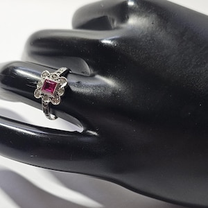 May include: A silver ring featuring a square red gemstone surrounded by smaller clear gemstones.