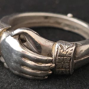 May include: A silver-toned ring featuring two clasped hands, symbolizing unity. The ring has decorative floral and geometric patterns on the band. The hands are detailed with visible fingers and a smooth, polished finish. The ring is set against a dark background.