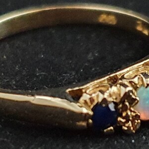 K&ouml;nnte beinhalten: Ein goldener Ring mit drei Edelsteinen. Der mittlere Stein ist ein wei&szlig;er Opal, und die beiden Steine an den Seiten sind blaue Saphire.