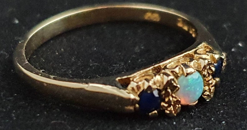 K&ouml;nnte beinhalten: Ein goldener Ring mit drei Edelsteinen. Der mittlere Stein ist ein wei&szlig;er Opal, und die beiden Steine an den Seiten sind blaue Saphire.