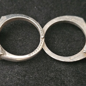 May include: A silver-toned bracelet with two connected circular bands. Each band features a hand-shaped clasp and decorative detailing. The bracelet is displayed on a dark surface.