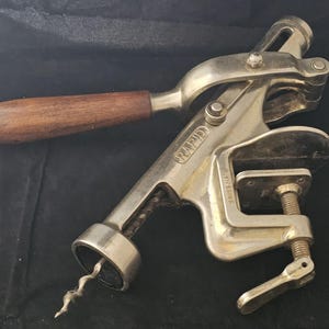 May include: A vintage Rapid corkscrew with a wooden handle and a silver-coloured metal body. The corkscrew has a clamp for securing it to a surface. The text "RAPID" and "SWISS MADE" are visible.