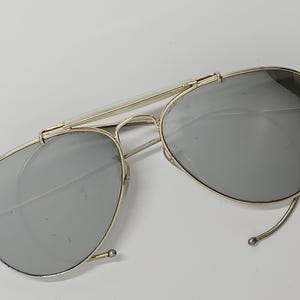 Pu&ograve; includere: Occhiali da sole in stile aviatore con montatura in metallo color oro e lenti grigie. Gli occhiali hanno un doppio ponte e una classica forma a goccia. Le lenti sono riflettenti.