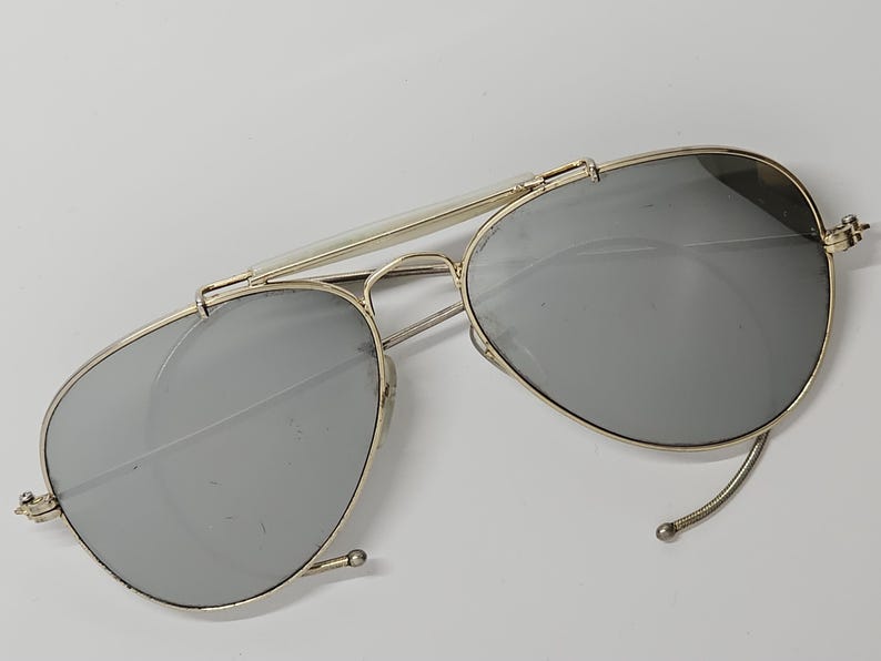 Pu&ograve; includere: Occhiali da sole in stile aviatore con montatura in metallo color oro e lenti grigie. Gli occhiali hanno un doppio ponte e una classica forma a goccia. Le lenti sono riflettenti.