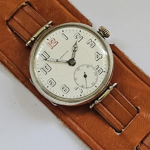 Op de afbeelding: Een zilveren Zenith polshorloge met een witte wijzerplaat en zwarte Arabische cijfers. Het horloge heeft een bruin leren bandje.