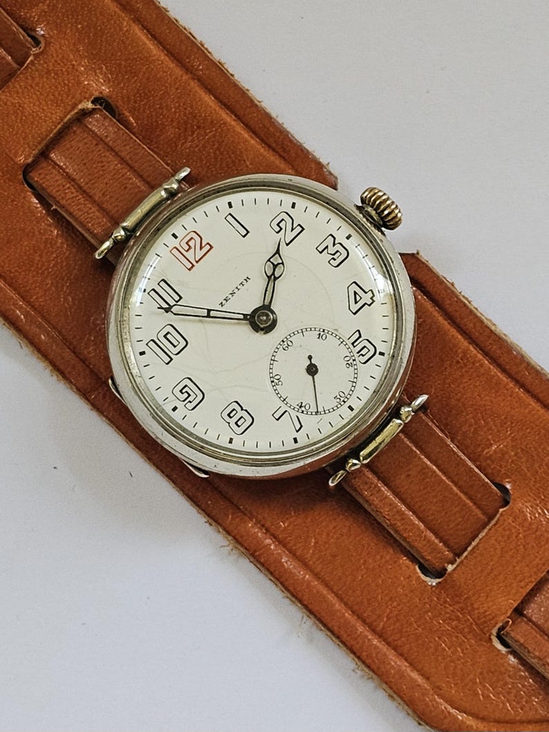 Op de afbeelding: Een zilveren Zenith polshorloge met een witte wijzerplaat en zwarte Arabische cijfers. Het horloge heeft een bruin leren bandje.