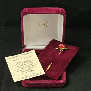 Può includere: Un spillo a forma di rosa, chiamato "La Rosa degli Amanti", in una scatola di velluto. Lo spillo è di colore rosso e oro e presenta una singola rosa rossa con foglie dorate e un diamante da 1/8 di punto. Il certificato di autenticità indica che lo spillo è stato realizzato in vermeil dorato e dipinto a mano con colori smaltati ricchi. Lo spillo è stato emesso in un'unica edizione rigorosamente limitata da The Franklin Mint.