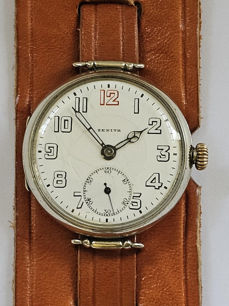 Op de afbeelding: Een zilveren Zenith polshorloge met een witte wijzerplaat en zwarte wijzers. Het horloge heeft een kleine secondewijzer-wijzerplaat onderaan de wijzerplaat. Het horloge zit op een bruin leren bandje.