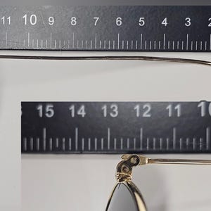 Pu&ograve; includere: Primo piano di occhiali con montatura argentata e lenti scure. Gli occhiali sono misurati contro un righello, mostrando una lunghezza di circa 14 cm. Il design presenta un'asta sottile e curva.