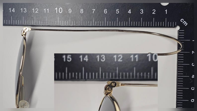 Pu&ograve; includere: Primo piano di occhiali con montatura argentata e lenti scure. Gli occhiali sono misurati contro un righello, mostrando una lunghezza di circa 14 cm. Il design presenta un'asta sottile e curva.