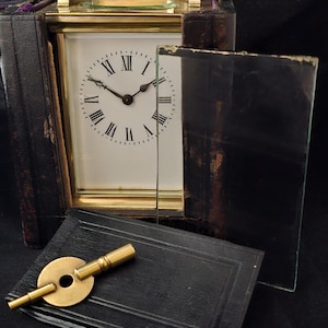 May include: An antique brass and glass clock with a white face and black Roman numerals. The clock is in a black leather case, with a brass key and a black leather-covered box. The clock face is visible through a glass panel.