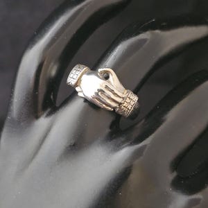 May include: A silver ring in the shape of two clasped hands. The ring has detailed patterns on the cuffs and the top of the ring. The ring is displayed on a black surface.