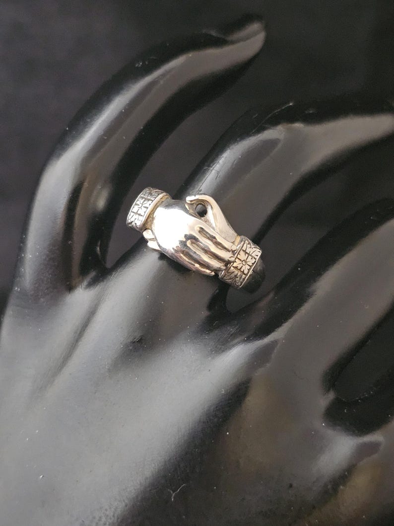 May include: A silver ring in the shape of two clasped hands. The ring has detailed patterns on the cuffs and the top of the ring. The ring is displayed on a black surface.